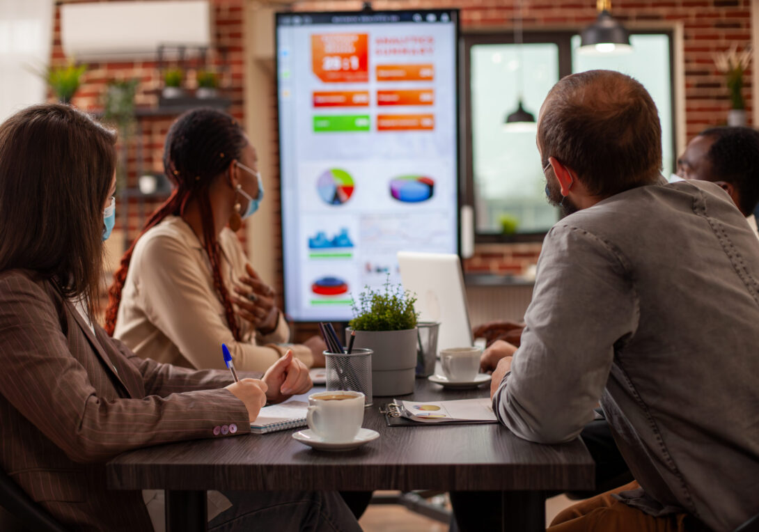 Coworkers seated at table with notepads, taking notes and discussing business project analytics shown on digital monitor. Professional team analyzing financial charts on led screen in startup office